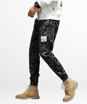 Side view of a man casually posing in black and grey camo cargo pants with eye-catching tan boots, modern street fashion.