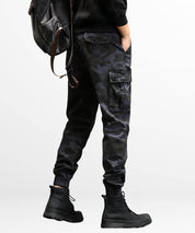 Rear view of dark grey camo cargo slim-fit pants on a man, highlighting the detailed back pocket and overall fit with a stylish leather backpack.