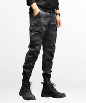 Side view of a man in dark grey camo cargo slim-fit pants showing utility pockets and tapered leg fit, paired with high-top black boots.