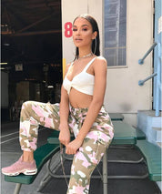 Casual look with woman in pink camouflage pants and white crop top, seated pose, outdoor urban setting.