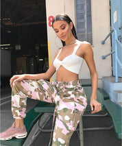 Trendy woman in pink camouflage pants and white crop top, relaxed pose with urban background, chic fashion look.