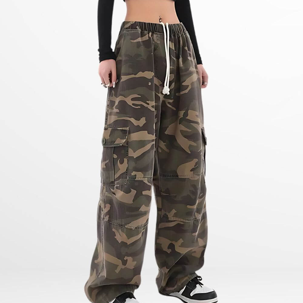 A front-facing view of a woman in streetwear style, wearing baggy camo pants with drawstrings and classic black and white sneakers.